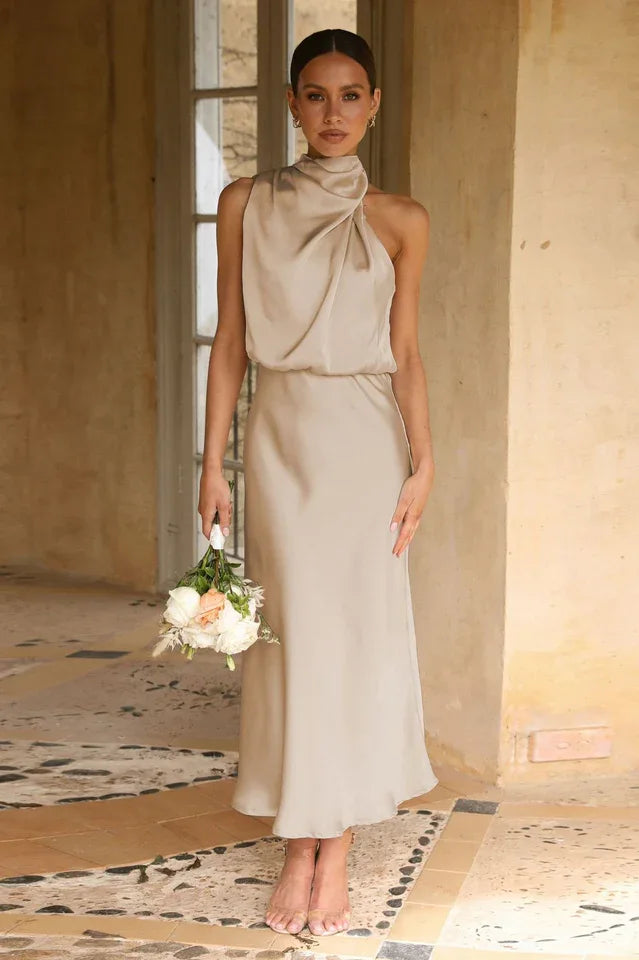 Woman wearing a champagne satin halter neck midi dress, holding a bouquet of flowers, standing near a door frame.