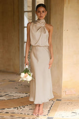 Woman wearing a champagne satin halter neck midi dress, holding a bouquet of flowers, standing near a door frame.