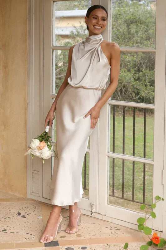 Woman wearing a champagne satin halter neck midi dress, holding a bouquet of flowers, leaning against a door frame.