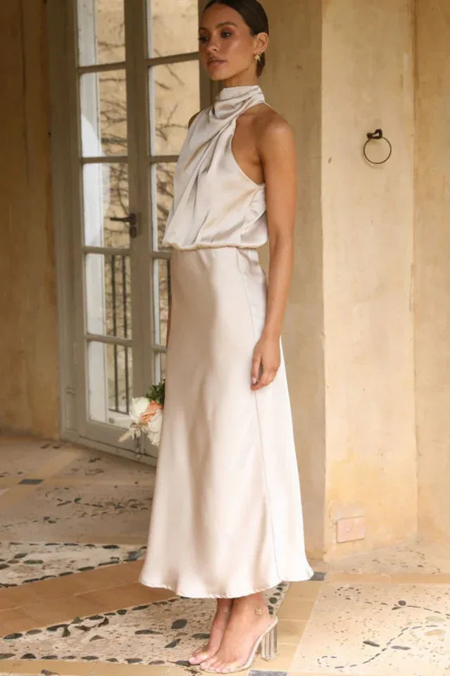 Side view of a woman wearing a champagne satin halter neck midi dress, standing near a door frame.