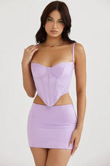 Model wearing a lavender corset top and skirt, hand on chest
