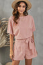 Woman wearing a pink romper with dolman sleeves and a tie waist, accessorized with a straw hat and gold jewelry.