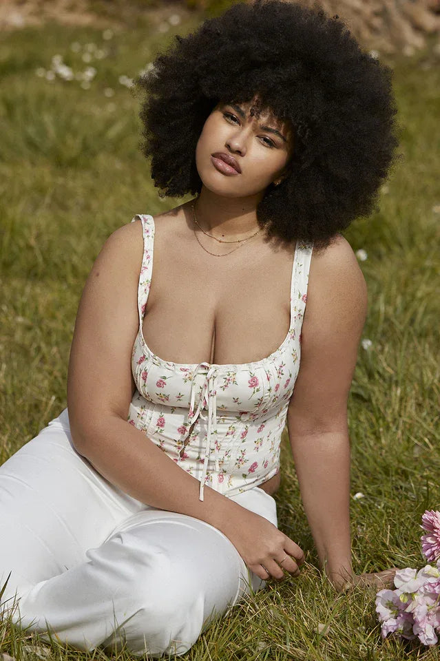Woman with an afro hairstyle wearing a white floral print corset top and white pants, sitting on grass.