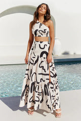 Woman wearing a cream and black abstract print one-shoulder crop top and a matching tiered maxi skirt with a high slit, standing by a pool.