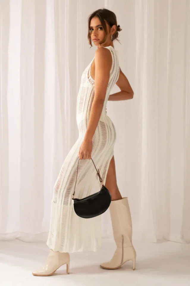 Side view of a woman in a white sleeveless maxi dress with a high slit and sheer knit, holding a black handbag, with cream boots.