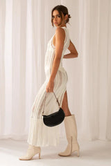 Side view of a woman in a white sleeveless maxi dress with a high slit and sheer knit, holding a black handbag, with cream boots.