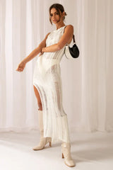 Woman in a white sleeveless maxi dress with a high slit and sheer knit, holding a black and white handbag, with cream boots.