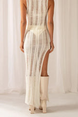 Back view of a woman wearing a white sleeveless maxi dress with a high slit and sheer knit construction, with cream boots.