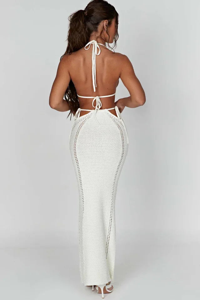 Back view of a woman wearing a white crochet knit halter neck bikini top and a matching white crochet knit high-waisted maxi skirt with cut-out details and tie closures.