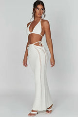 Side view of a woman in a white crochet knit halter neck bikini top and a matching white crochet knit high-waisted maxi skirt with cut-out details and tie closures, wearing white heeled sandals.