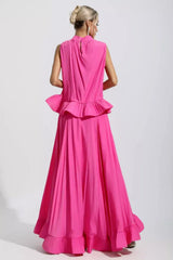 Back view of a woman wearing a bright pink sleeveless mock neck top with a ruffled peplum and a high-low maxi skirt.