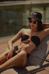 Woman in a black strapless bikini with gold accents and a black bucket hat, posing on a lounge chair.