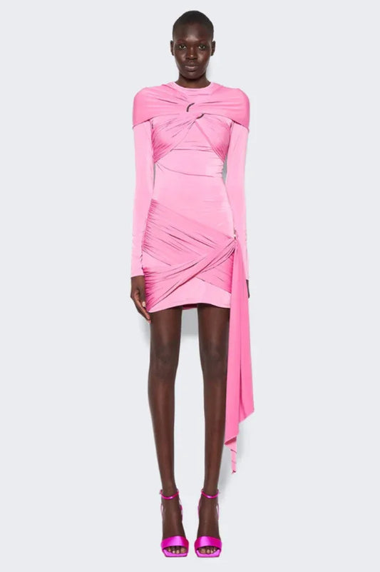Model wearing a pink long-sleeve mini dress with asymmetrical draping and a twist detail at the neckline. The dress has a cutout detail at the chest and a ring hardware detail on the hip. The hem is asymmetrical with a draped panel.