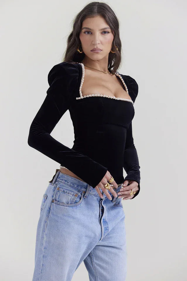 Side view of a model in a black velvet long-sleeve top with a square neckline and puffed shoulders, paired with light-wash denim jeans.