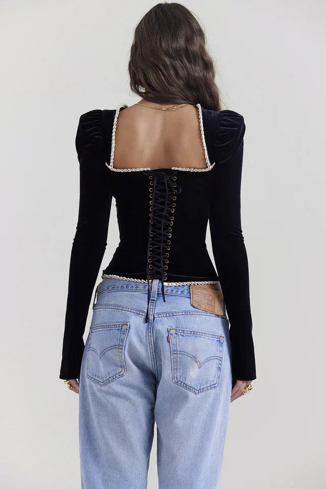 Back view of a model wearing a black velvet long-sleeve top with corset-style lacing, paired with light-wash denim jeans.
