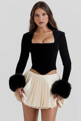 Woman wearing a black velvet long-sleeved top with faux fur cuffs and a cream pleated mini skirt.
