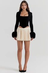 Full body shot of a woman wearing a black velvet long-sleeved top with faux fur cuffs and a cream pleated mini skirt, paired with black heels.