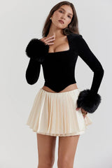 Side view of a woman in a black velvet top with faux fur cuffs and a cream pleated mini skirt.