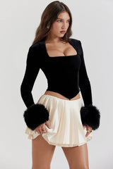 Close-up of a woman's torso wearing a black velvet top with faux fur cuffs and a cream pleated mini skirt.