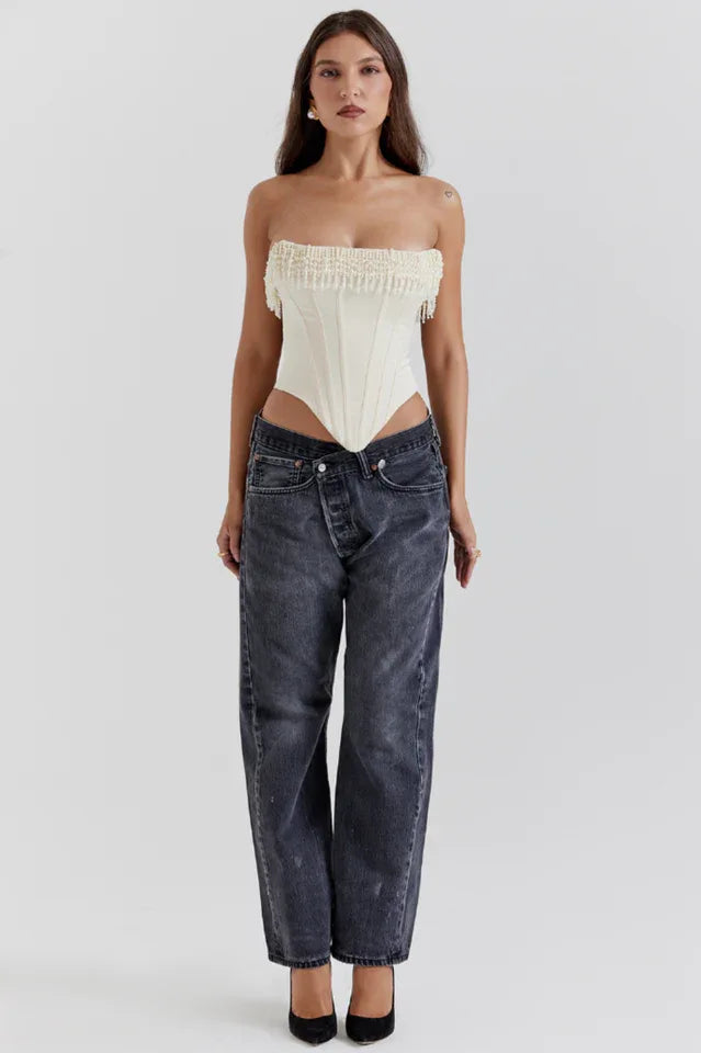 Full-body front view of a model wearing a cream strapless corset top with beaded fringe detail and dark wash jeans, paired with black heels.
