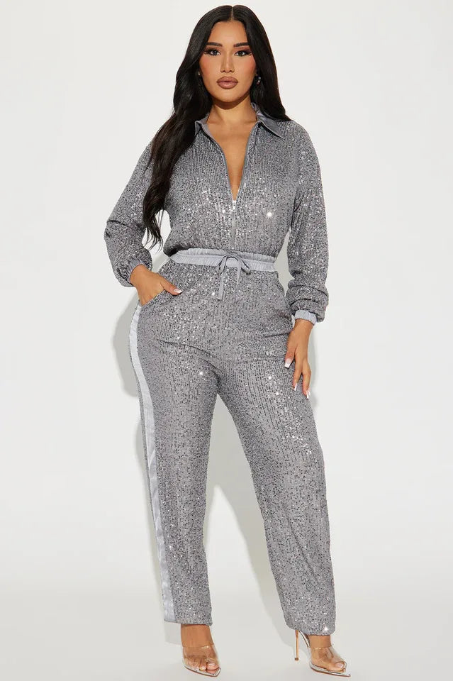 Front view of a woman wearing a grey sequined long-sleeved collared jumpsuit with a zipper front closure and drawstring waist. The pants have a grey side stripe.