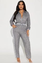 Front view of a woman wearing a grey sequined long-sleeved collared jumpsuit with a zipper front closure and drawstring waist. The pants have a grey side stripe.