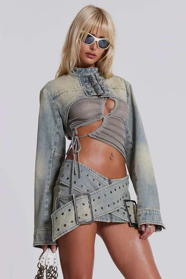 Woman wearing a light wash distressed denim cropped jacket over a grey ruched top and a matching mini skirt with buckle detailing, accessorized with sunglasses.