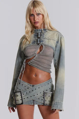 Woman wearing a light wash distressed denim cropped jacket over a grey ruched top, paired with a matching mini skirt with buckle detailing.