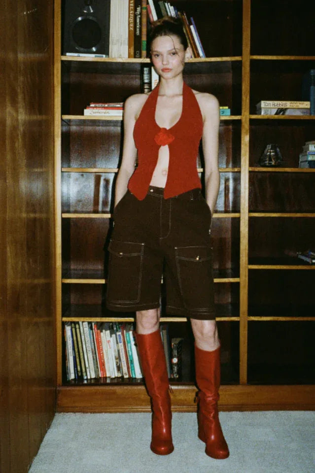 Model wearing a red halter top, brown shorts, and red boots standing in front of a bookshelf.