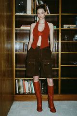 Model wearing a red halter top, brown shorts, and red boots standing in front of a bookshelf.