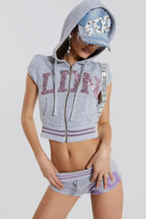 Model wearing a gray cropped zip-up hoodie with short sleeves and sequin embellishments on the front, accessorized with a denim cap and sunglasses, hands placed on hips.