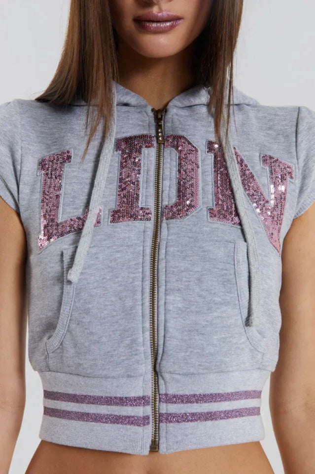 Close-up of the front of a gray cropped zip-up hoodie with short sleeves, featuring sequin embellishments on the chest and striped trim at the hem.