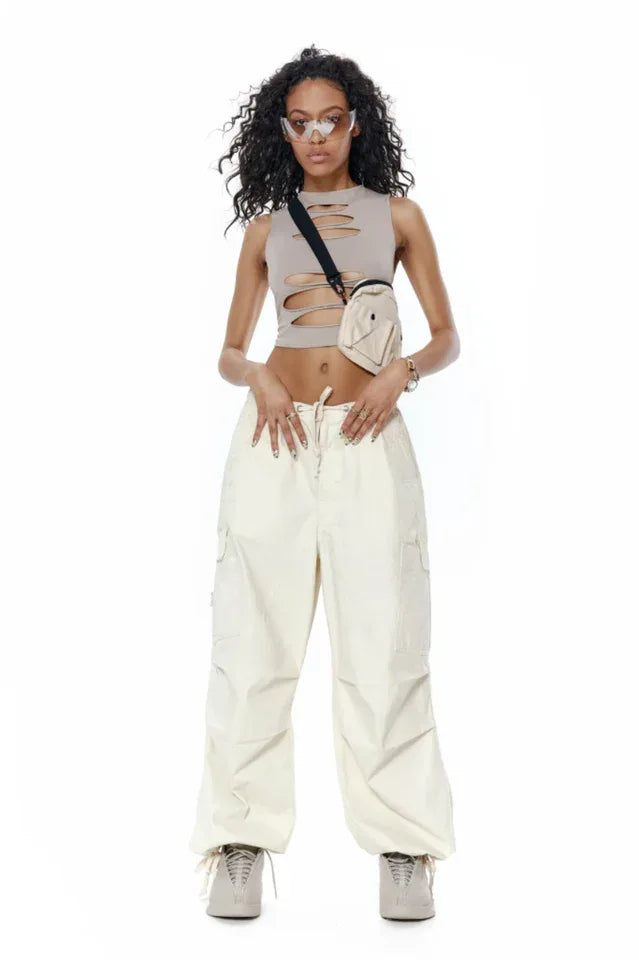 Full-body shot of a model wearing a taupe sleeveless cropped top with cut-outs, light-colored baggy cargo pants, and sneakers.
