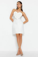 Woman wearing a white strapless mini dress with a heart-shaped applique on the bust, standing with her legs crossed.