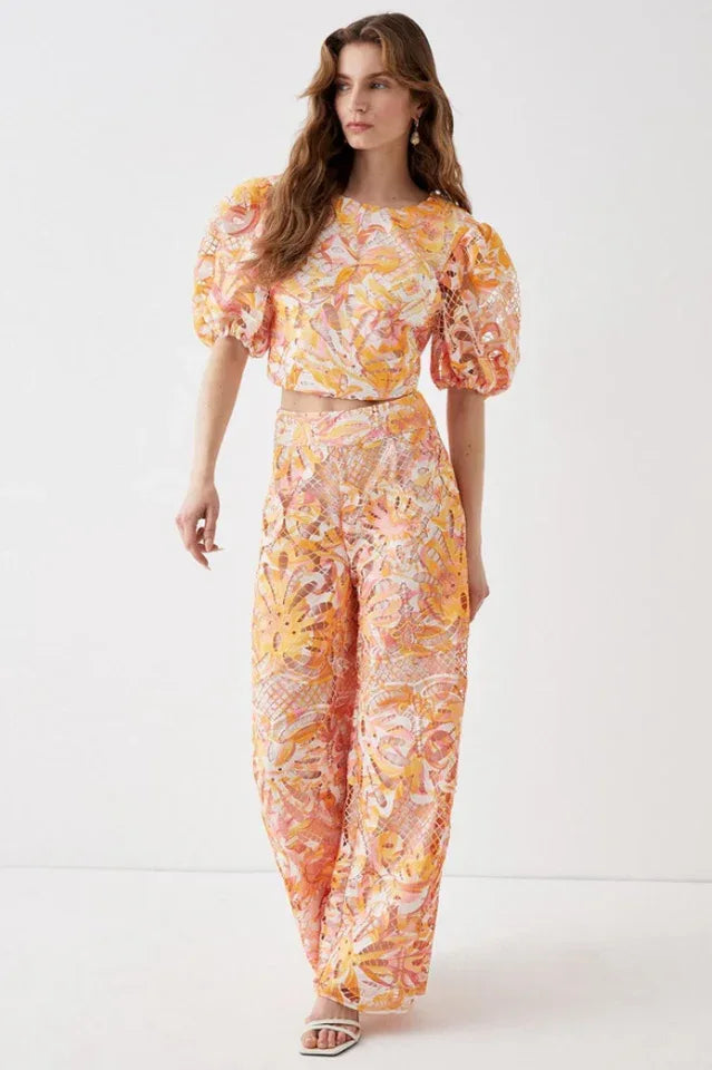 Model wearing a two-piece set with a cropped top and wide-leg pants featuring a floral lace overlay in orange, pink, and yellow.