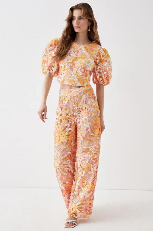 Model wearing a two-piece set with a cropped top and wide-leg pants featuring a floral lace overlay in orange, pink, and yellow.