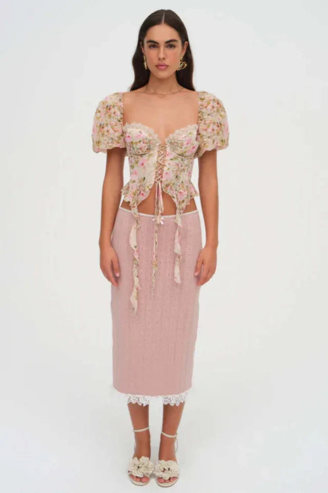 Full body shot of a woman wearing a floral print corset top with puffy sleeves and a lace-up front, paired with a pink skirt and sandals.