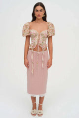 Full body shot of a woman wearing a floral print corset top with puffy sleeves and a lace-up front, paired with a pink skirt and sandals.