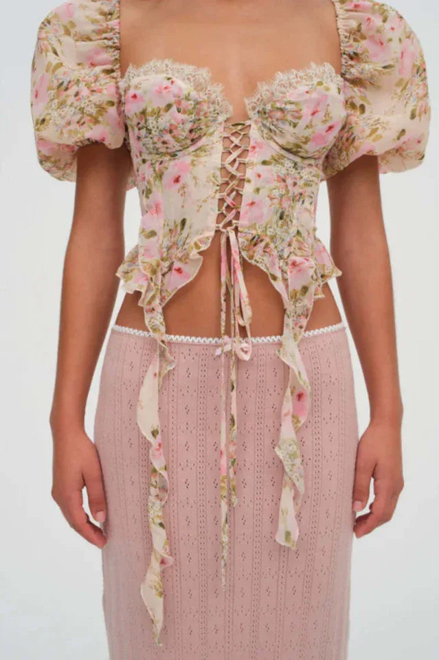 Front view of a woman wearing a floral print corset top with puffy sleeves and a lace-up front, paired with a pink skirt.