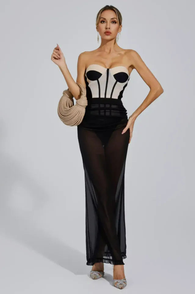 Woman in a strapless corset dress with a sheer black maxi skirt, holding a beige woven handbag and gesturing with her other hand.