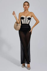 Woman in a strapless corset dress with a sheer black maxi skirt, holding a beige woven handbag and gesturing with her other hand.