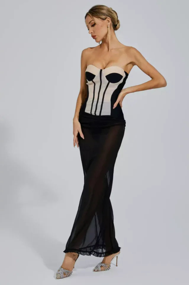 Woman in a strapless corset dress with a sheer black maxi skirt, looking down with one hand on her hip.