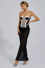 Woman in a strapless corset dress with a sheer black maxi skirt, looking down with one hand on her hip.
