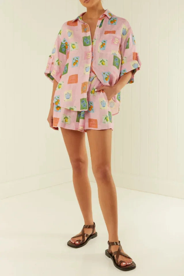 Woman standing in a room wearing a pink printed linen blend matching set with a camp collar shirt and belted shorts, paired with brown strappy sandals.