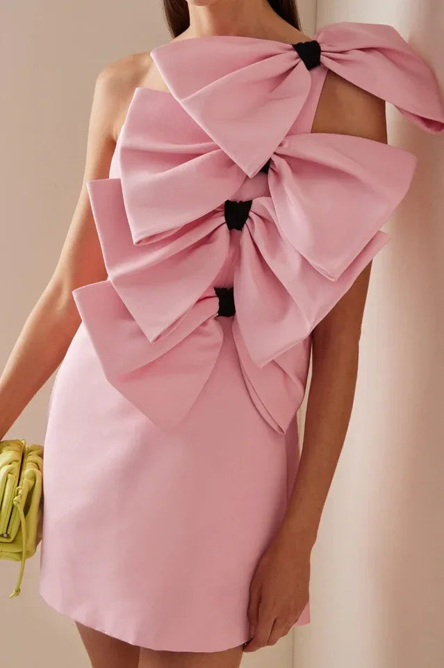 Pink one-shoulder mini dress with large bow embellishments on the bodice and shoulder, held by a person with a lime green clutch bag.