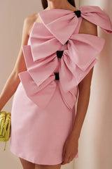 Pink one-shoulder mini dress with large bow embellishments on the bodice and shoulder, held by a person with a lime green clutch bag.