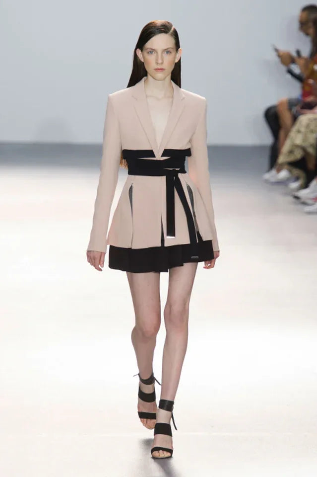 Full runway shot of a model wearing a light pink blazer with a black wrap belt and black skirt.