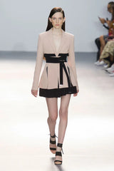 Full runway shot of a model wearing a light pink blazer with a black wrap belt and black skirt.
