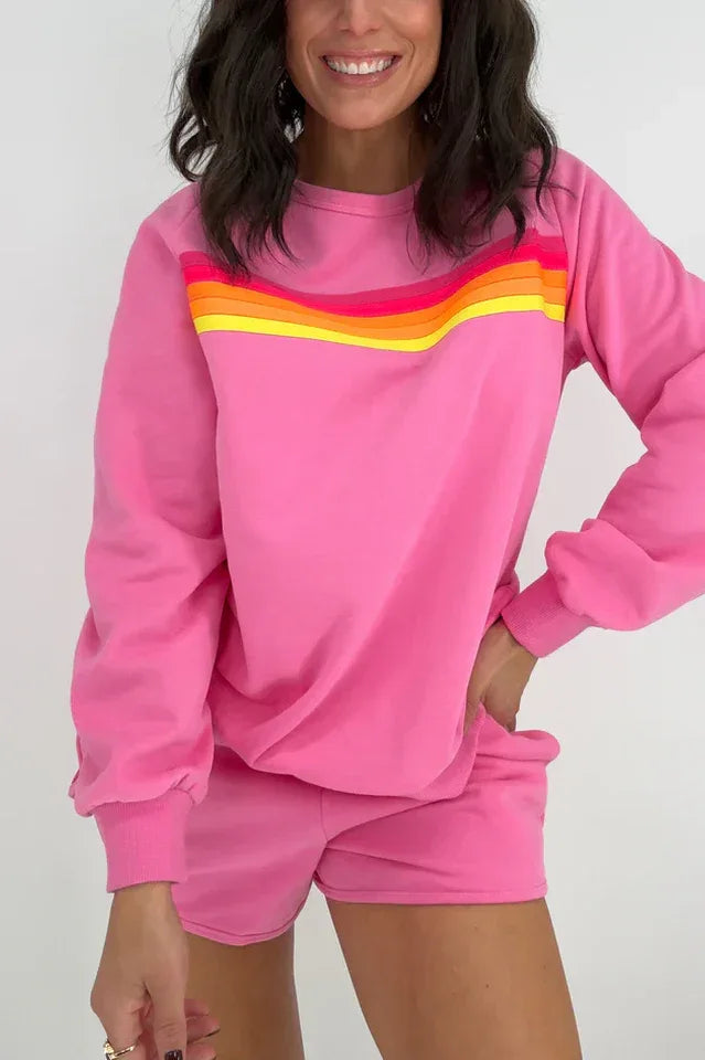 Woman wearing a pink crew neck sweatshirt with rainbow stripes across the chest and matching pink shorts with rainbow stripes on the side.