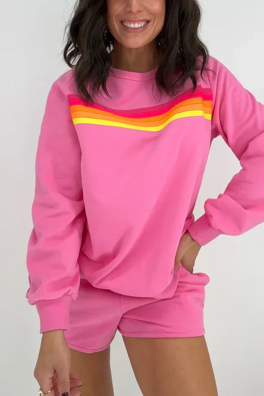Woman wearing a pink crew neck sweatshirt with rainbow stripes across the chest and matching pink shorts with rainbow stripes on the side.
